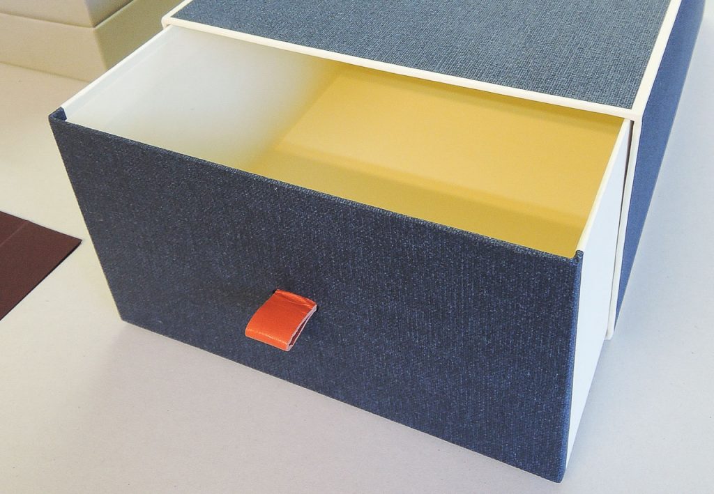 Geöffnete Schiebeschachtel, innen cremeweiss, aussen mit graublauem Bezugspapier und orangefarbener Grifflasche aus Leder. 