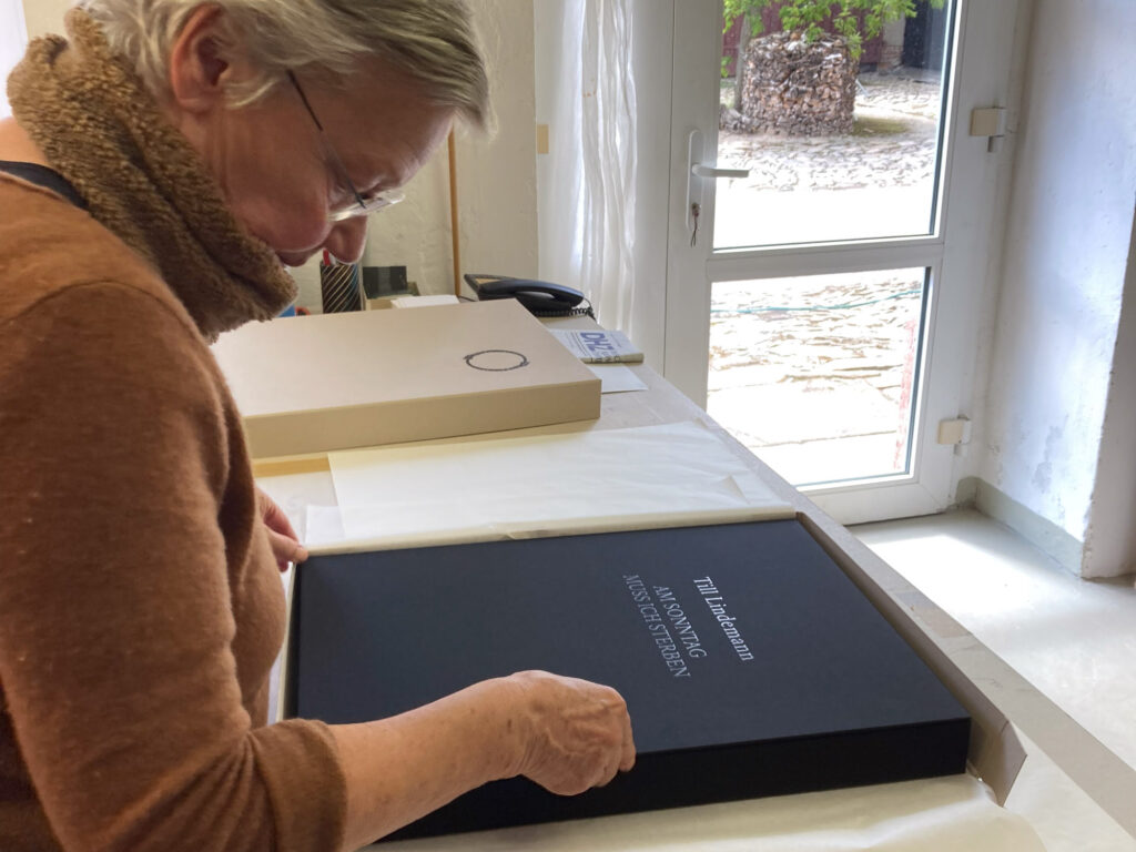 Im Vordergrund die Verpackung einer Kunstbuchkassette des Gedichtbandes Till Lindemann "Am Sonntag muss ich sterben", mit schwarzem Leinenband und geprägtem Titel, in die entsprechende Transportverpackung aus Graupappe. Im Hintergrund der ebenfalls geprägte Deckel der zugehörigen Transportverpackung.