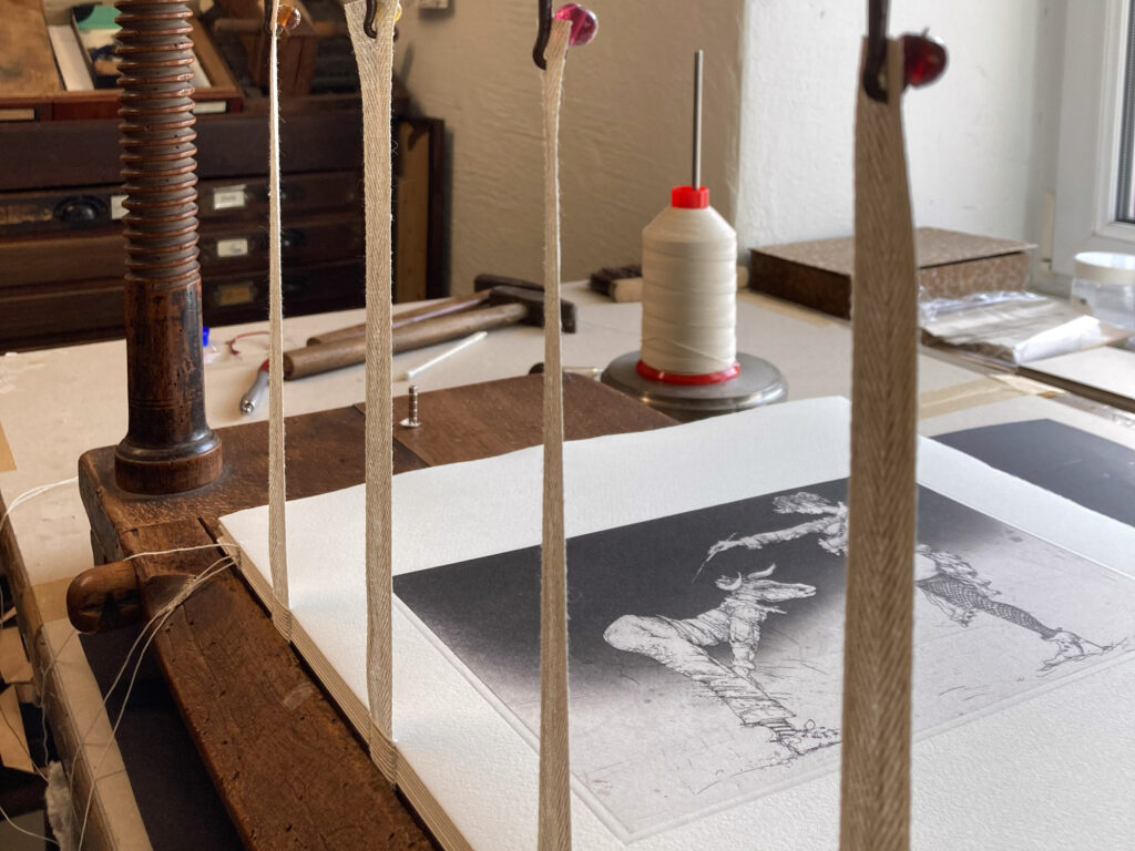 Im Vordergrund eine von Bodo Klös illustrierte Buchseite des Gedichtbandes "Am Sonntag muss ich sterben" von Till Lindemann, in einer Heftlade. Im Hintergrund der Arbeitstisch mit Werkzeugen.